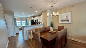 Uma área de refeições com mesa de madeira e cadeiras de vime, cozinha ao fundo e um lustre.