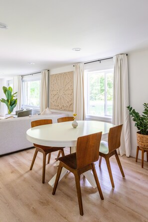 A dining area with a round table, wooden chairs, and a white tablecloth. There is a large window with curtains, a plant, and a decorative wall piece.