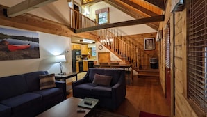 A cozy living room with a sofa, coffee table, and a painting of a red canoe on a lake. A staircase leads to a second floor with a wooden railing. - Wilderness Presidential Resort Cabins (Spotsylvania)