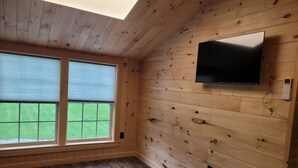 Ein Zimmer mit Holzwänden, einem großen Fenster mit Jalousien, einem an der Wand montierten Flachbildfernseher und einem Holzboden.