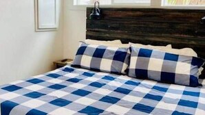 A bedroom with a checkered bedspread, a wooden headboard, a nightstand, and a window with a view of greenery.