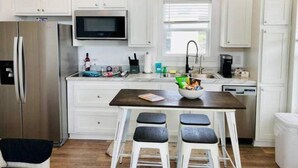 A kitchen with white cabinets, a stainless steel refrigerator, a microwave, a sink, and a coffee maker. There are three stools and a small table with a bowl of snacks.