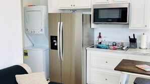 A compact kitchen with white cabinets, a stainless steel refrigerator, a microwave, and a small dining area with a table and chairs. - The Spoonbill Guest House at Goose Island Flats 1BR 1BA (Rockport)