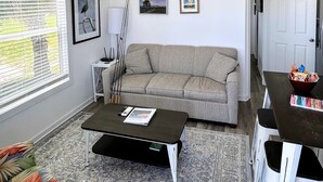 A modern living room with a grey sofa, a black coffee table, a ceiling fan, and a view of the outdoors through large windows. - The Spoonbill Guest House at Goose Island Flats 1BR 1BA (Rockport)