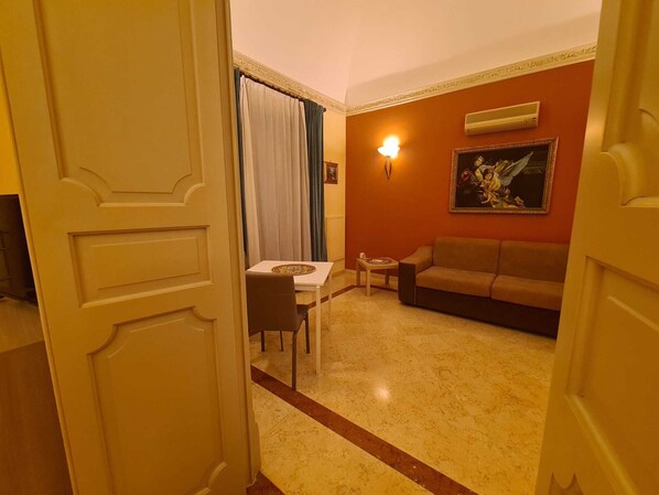 A hotel room with a dining table, a sofa, a painting on the wall, and an air conditioning unit.