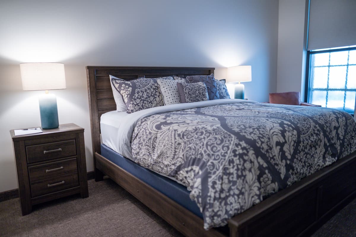 A bedroom with a wooden bed, patterned bedding, two bedside tables with lamps, and a window with blinds.