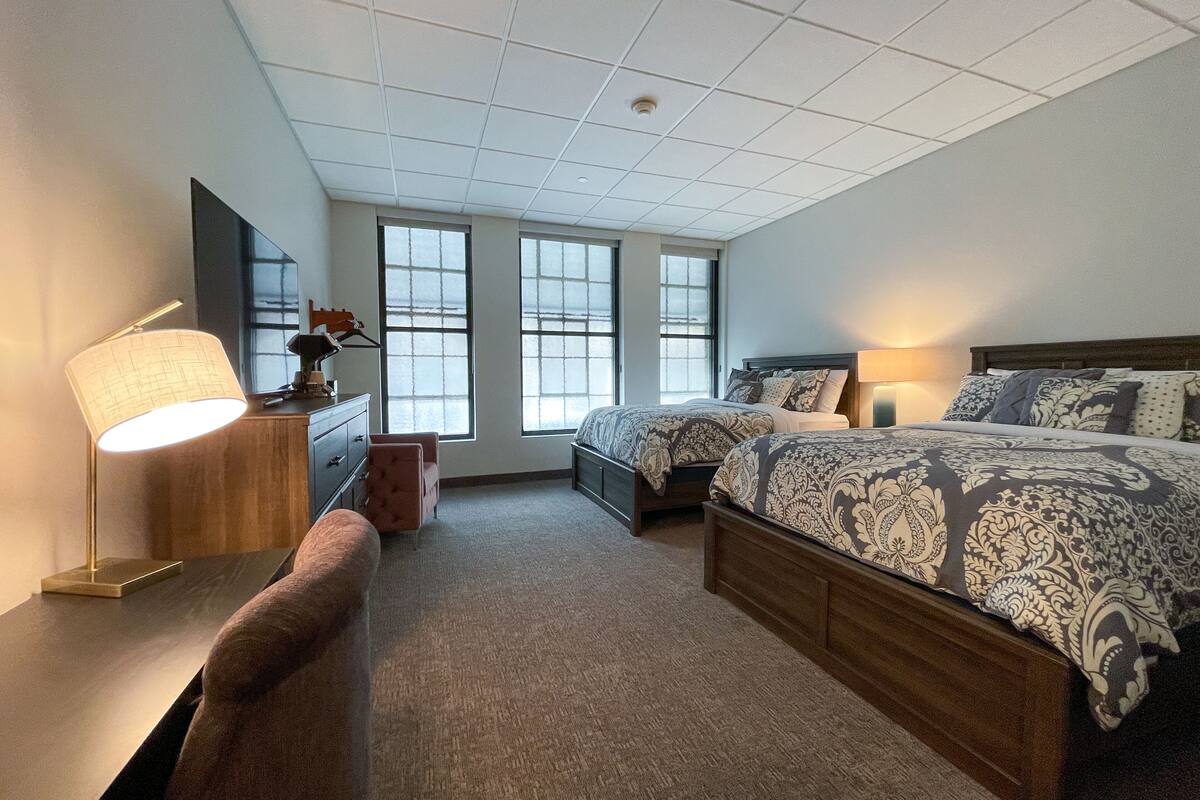 A bedroom with a large bed, a desk with a lamp, a chair, and a dresser.