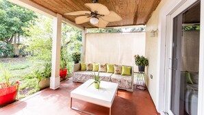 En overdækket terrasse med ventilator, et hvidt sofabord, en sofa med puder og krukkeplanter.