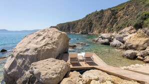 A wooden deck with two lounge chairs overlooking a clear blue sea and rocky coastline.