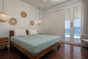 A bedroom with a wooden bed, a nightstand, a large window with a view of the sea, and three wall-mounted lamps.