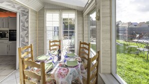 Un comedor con una mesa dispuesta para cuatro personas, rodeada de sillas de madera, bajo una estructura de vidrio con vista a un patio trasero.