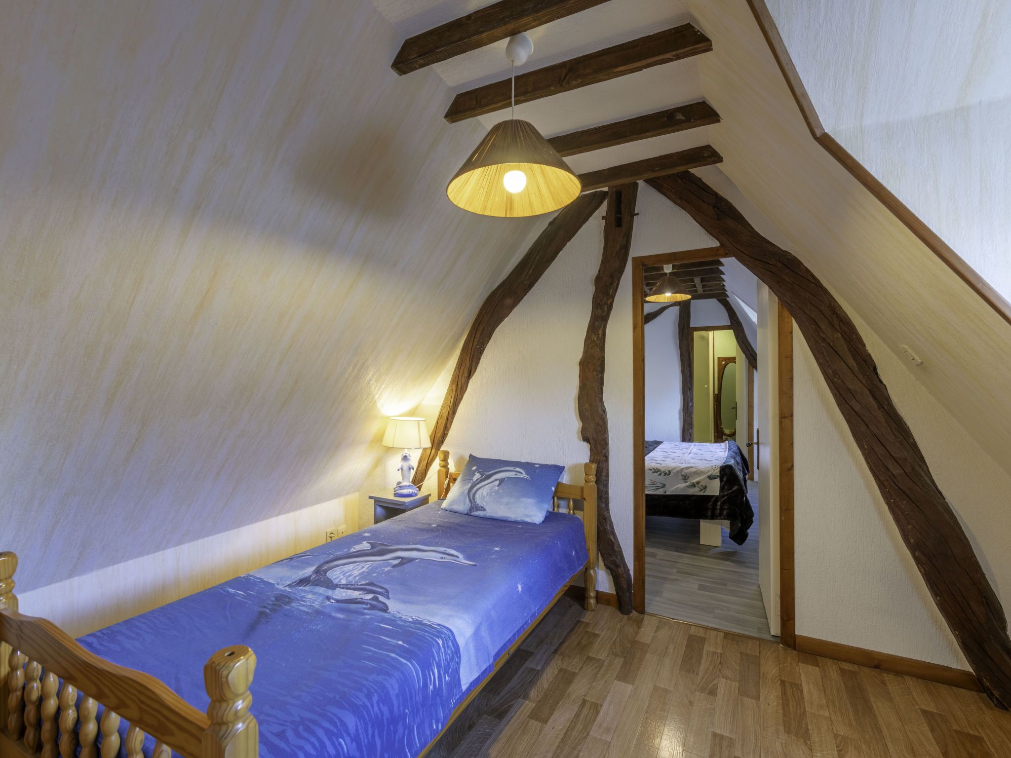Ein Schlafzimmer mit schrägem Dach, Holzbalken, einem Bett mit blauer Bettdecke, einer Hängelampe und einem Nachttisch mit Lampe.