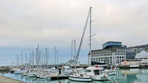 Marina con varios veleros amarrados, un edificio moderno con fachada de vidrio curvo y un paseo marítimo empedrado.