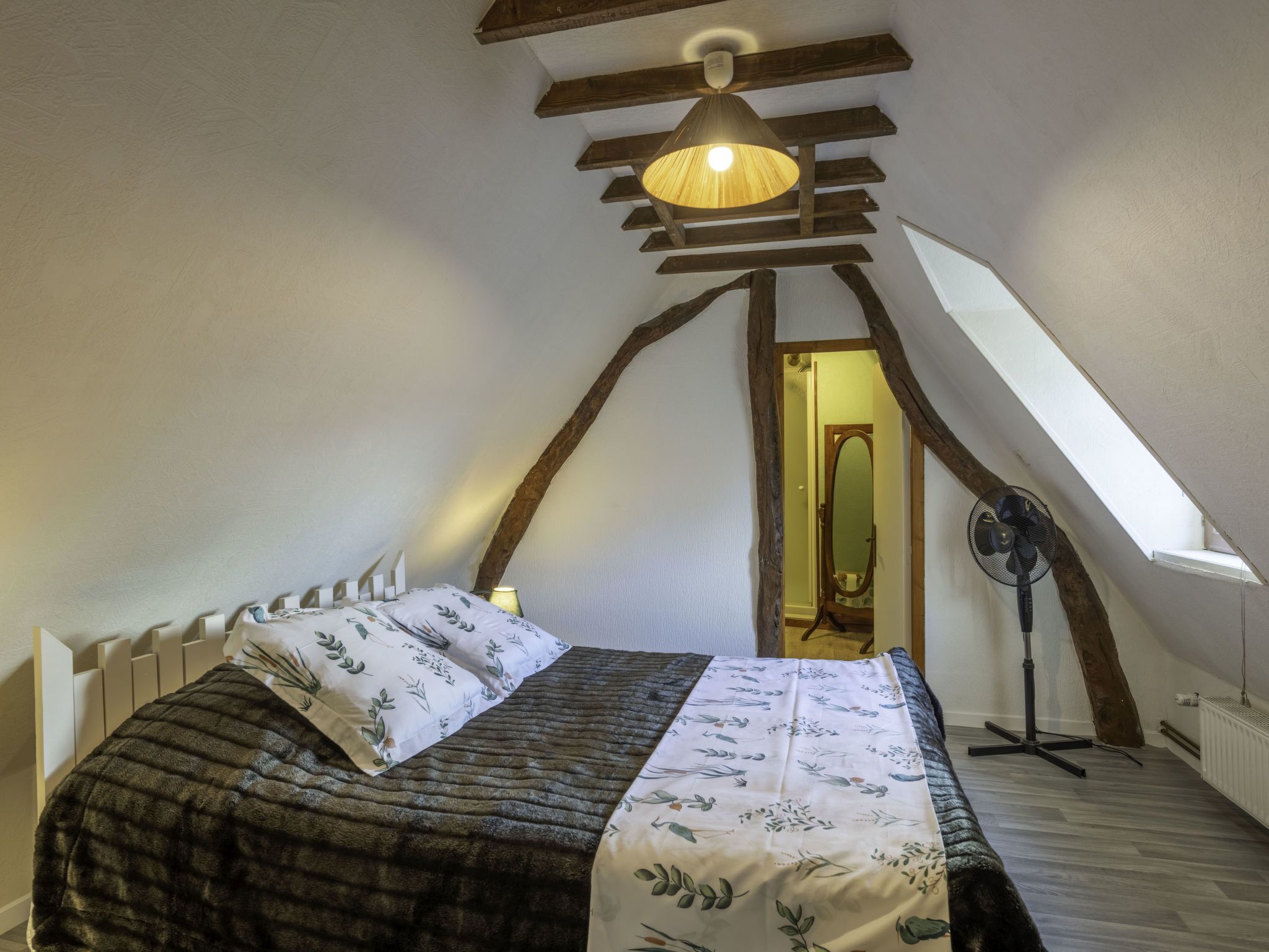 Une chambre avec un plafond en pente, un lit avec une literie à motifs floraux, un ventilateur sur pied et un puits de lumière.