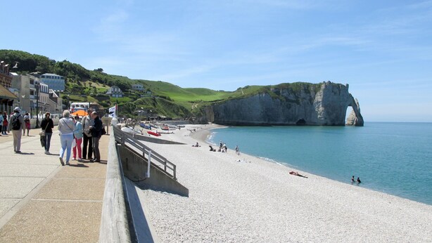 Un paseo marítimo junto a la costa, con una playa de guijarros, un acantilado con un arco y un pueblo con edificios en la ladera.