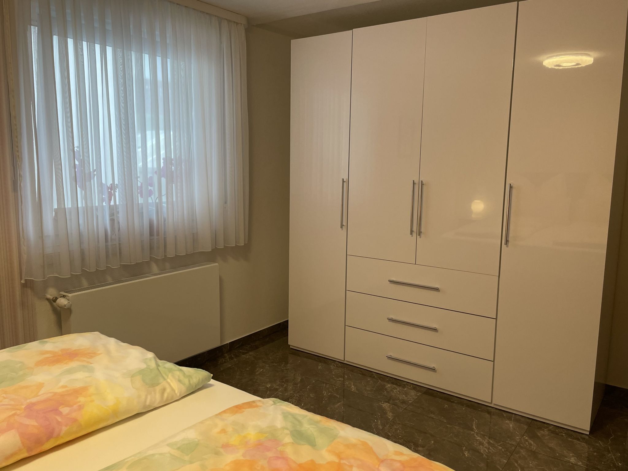 Een slaapkamer met een bed, een kledingkast en een raam met dunne gordijnen.