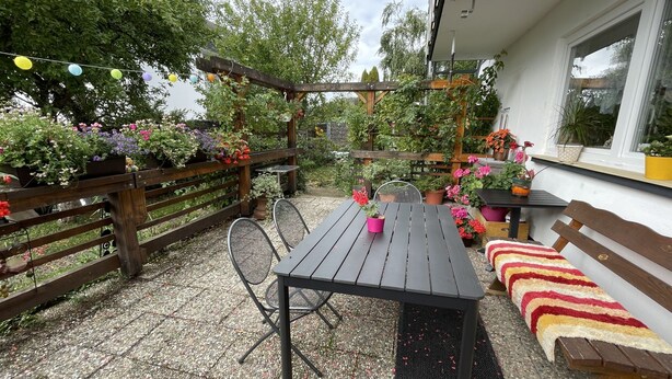 En terrasse med et træbord og stole, omkranset af et træhegn og krukker med planter.