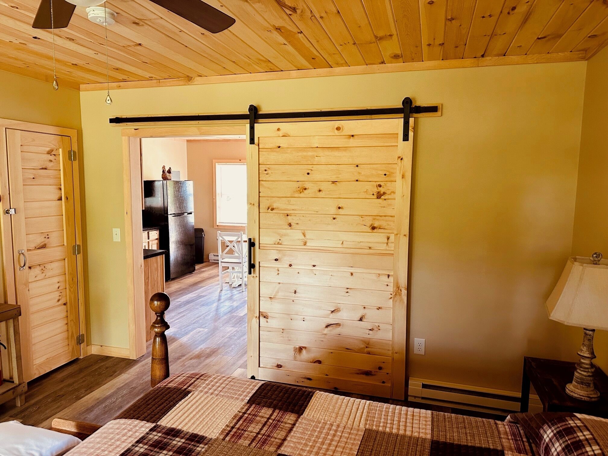 A room with a wooden floor, a bed with a plaid bedspread, a sliding door, a ceiling fan, and a kitchenette in the background.