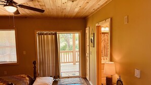 A room with a bed, a ceiling fan, a window with blinds, a door leading outside, and a small table with a lamp. - Cozy Cabin Boarding Winslow Hill Elk Viewing Area! (Benezette)