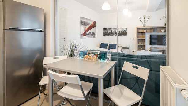 A modern kitchen with a stainless steel refrigerator, a dining table with two white chairs, and a sofa in the background. - Cozy spacious studio in new building (București)