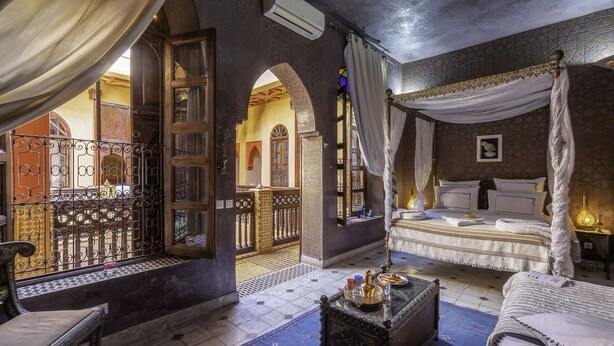 Ein traditioneller marokkanischer Raum mit einem Himmelbett, einem kleinen Tisch mit Dekoration und Blick auf den Innenhof durch eine offene Tür.