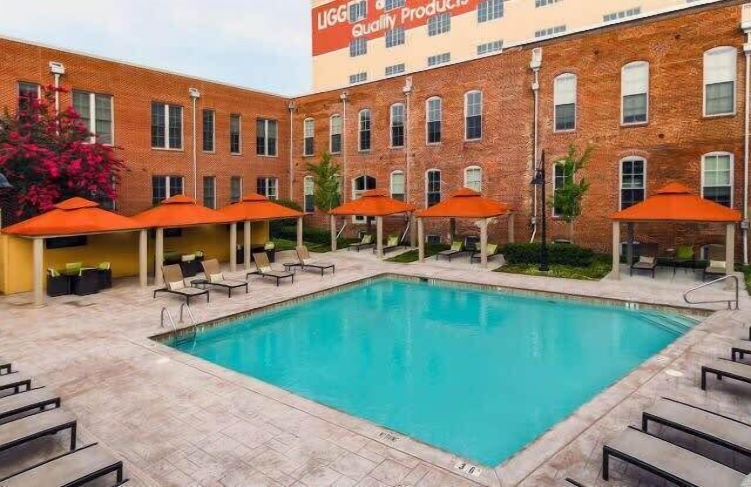 Una piscina con sillones de descanso y sombrillas, rodeada por un edificio de ladrillo con un letrero que dice "LIGG Quality Products".