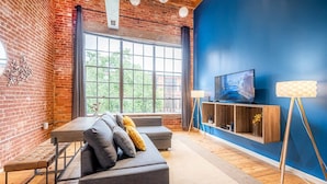 Un salon moderne avec un mur en briques, une grande fenêtre, un canapé gris, un plancher en bois et un téléviseur posé sur une étagère en bois.