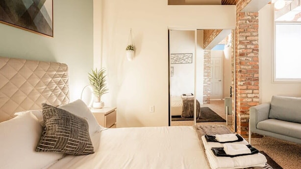 Une chambre avec un grand lit, une table de chevet ornée d’une plante, et une vue sur un mur de briques et une autre pièce.