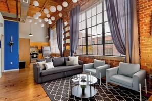 Una sala moderna con un sofá modular, dos sillones y una mesa de centro. Se ve un área de cocina con una estufa y un refrigerador al fondo.