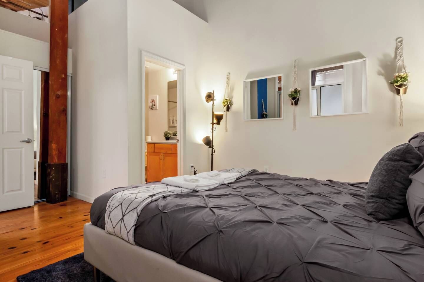 Une chambre avec un grand lit, un plancher en bois, une porte blanche et une petite fenêtre ornée d’une plante.