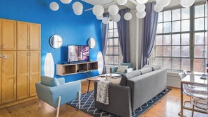 A modern living room with a blue accent wall, a grey sofa, a TV, and a dining area with a table and chairs. - 100 Year-Old Historic Brick 2BR Loft-High Ceiling8 (Durham)
