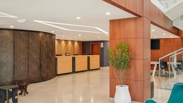 A modern hotel reception area with a wooden reception desk, a large potted plant, and a staircase.