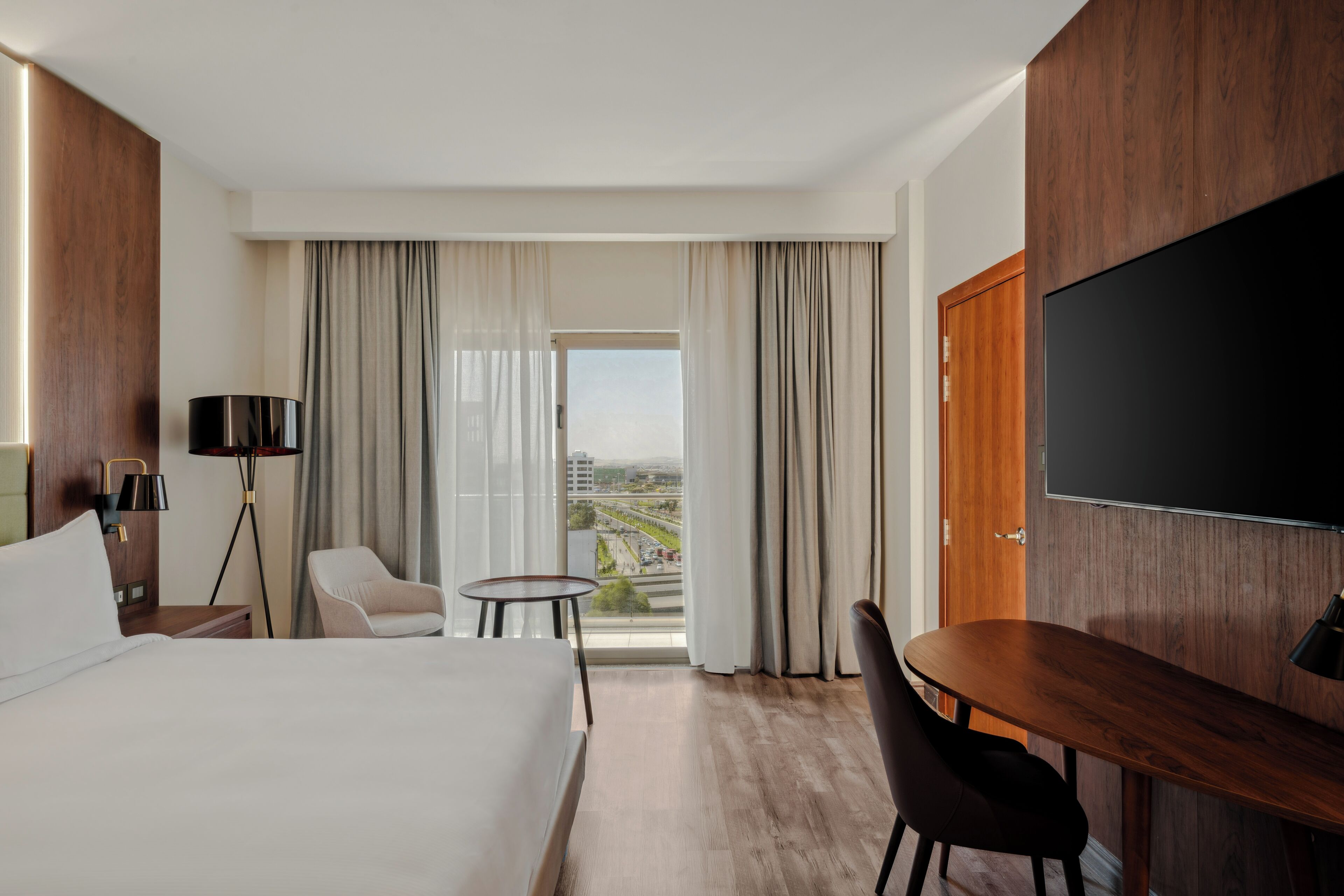 Une chambre d’hôtel avec un grand lit, un bureau, une chaise, une télévision et une vue sur la ville par la fenêtre.
