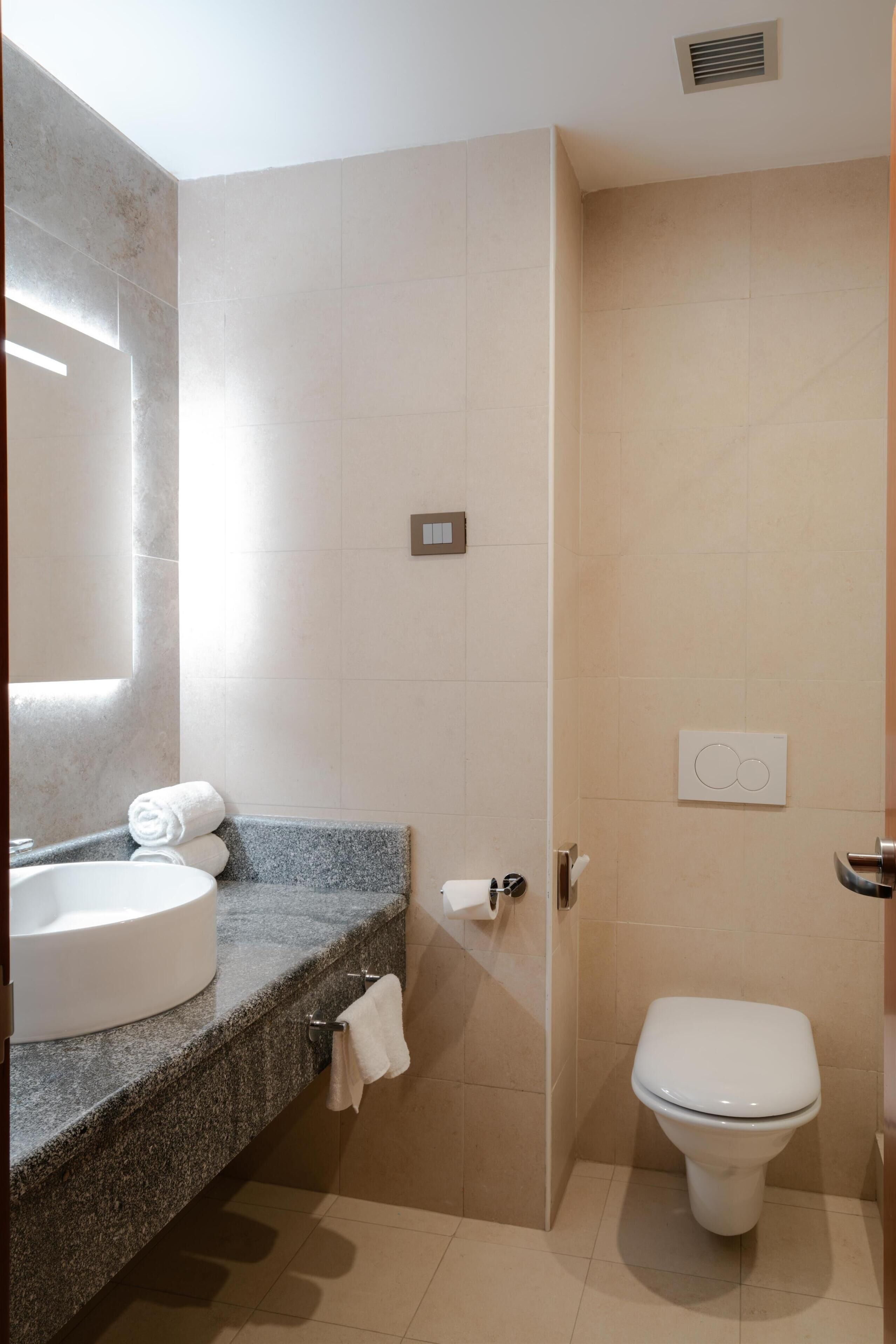 Un baño moderno con encimera de granito, lavabo redondo, inodoro y toallero.