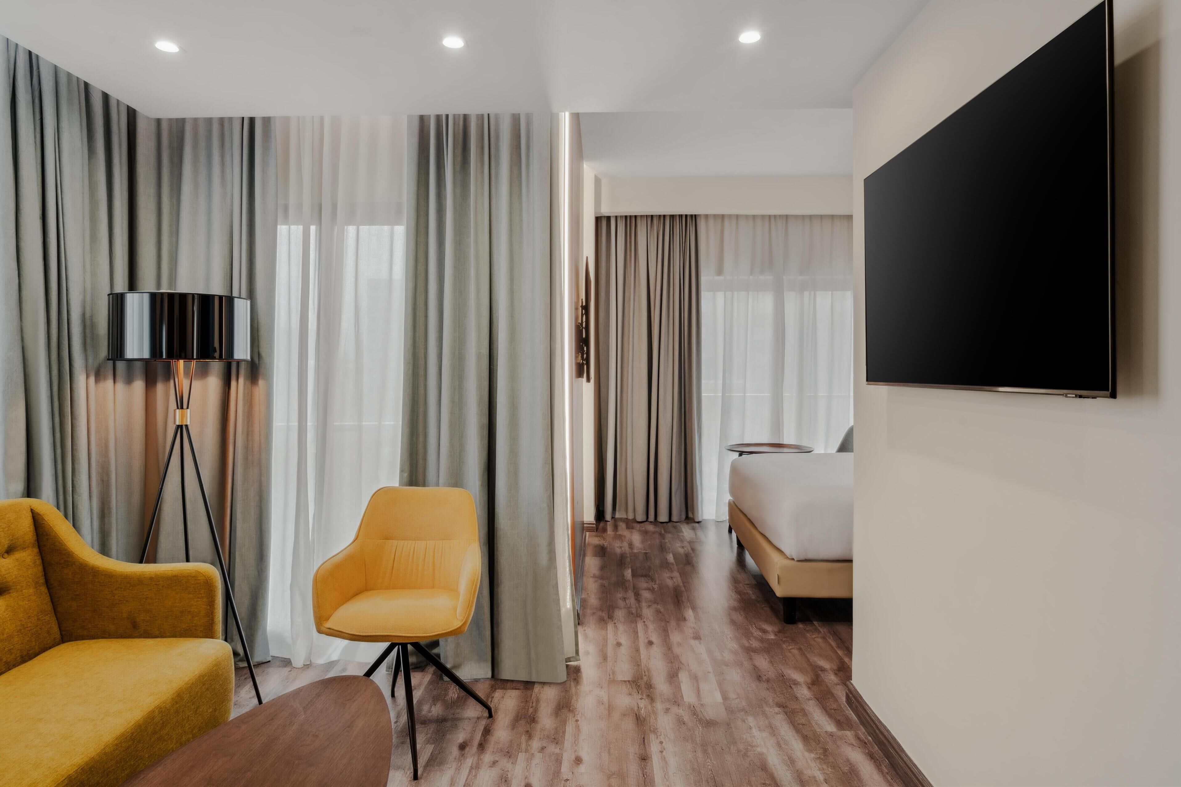 Een hotelkamer met een houten vloer, een bank, een stoel, een bed, een televisie en gordijnen.