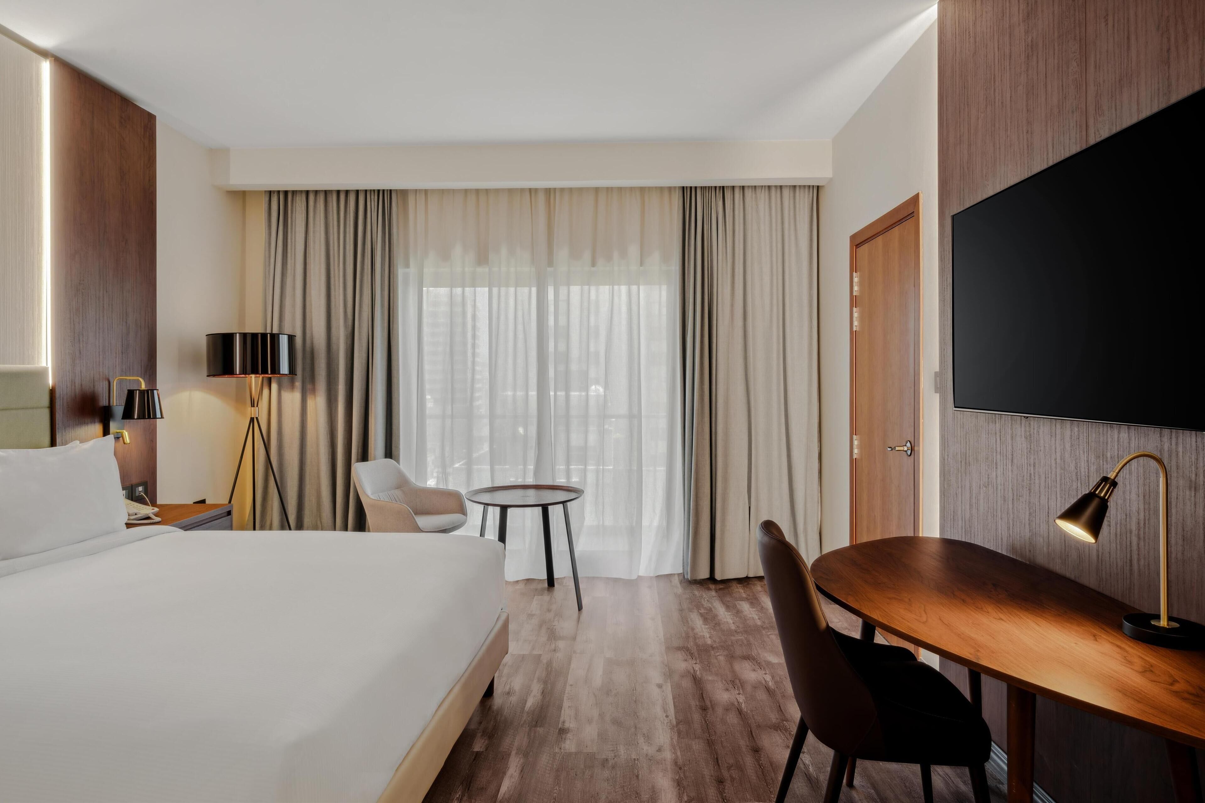 A hotel room with a large bed, a desk, a chair, a lamp, a TV, and a window with curtains.