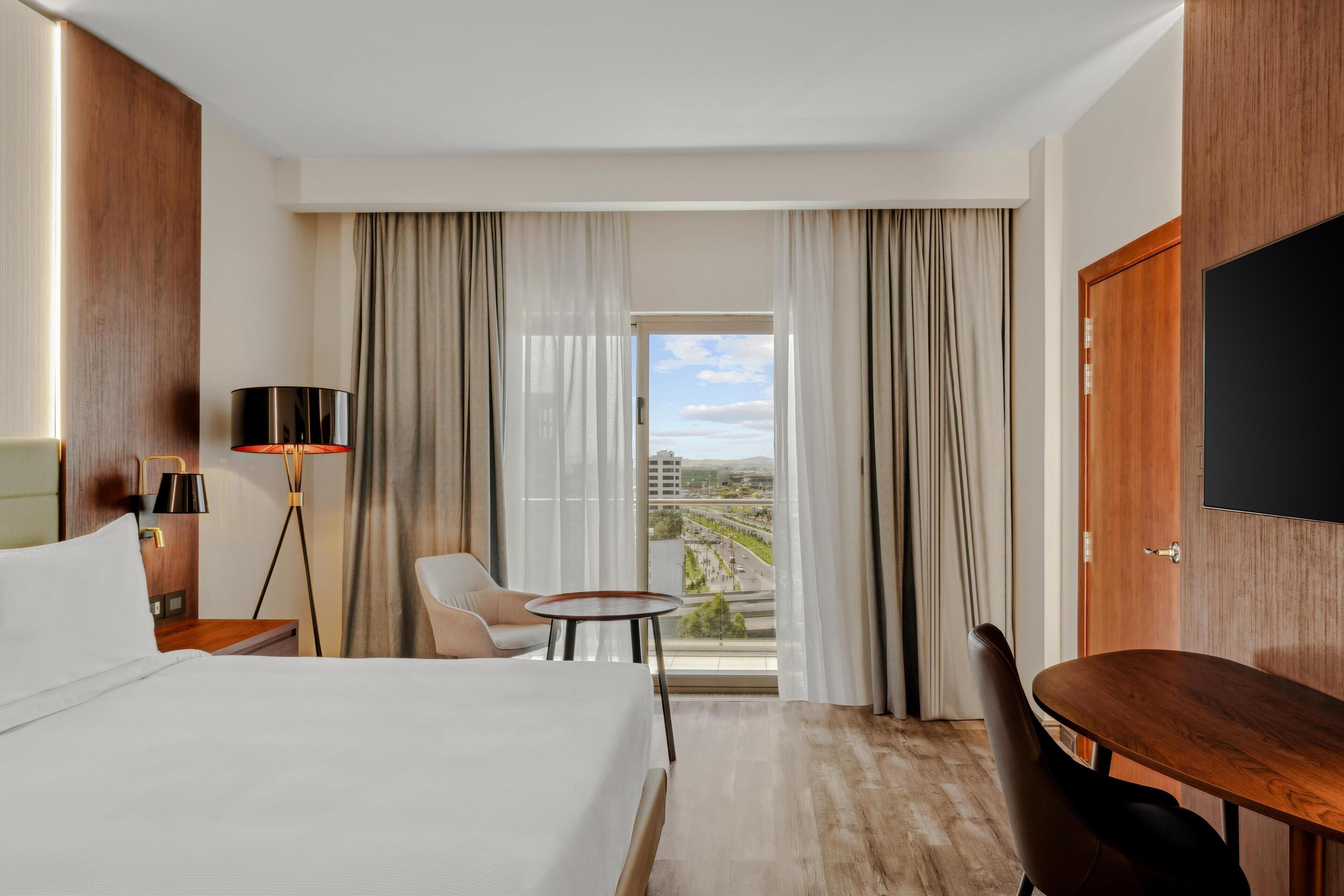 A hotel room with a large bed, a desk, a chair, a TV, and a view of the city through the window.