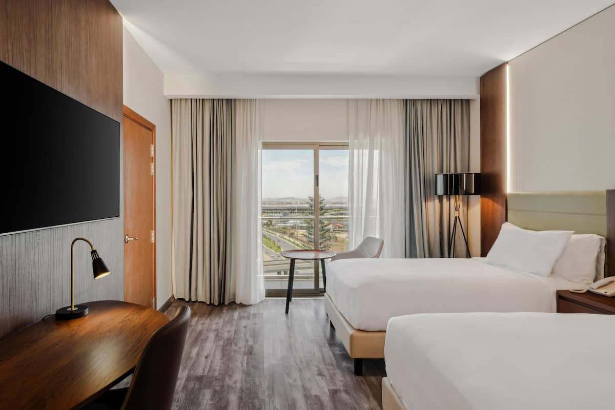 Une chambre d’hôtel avec deux lits, un bureau, une télévision et une grande fenêtre donnant sur la ville.