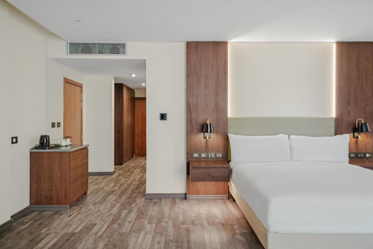 Une chambre d’hôtel avec un grand lit, une tête de lit en bois, des tables de chevet avec des lampes, une commode en bois et un petit coin cuisine.