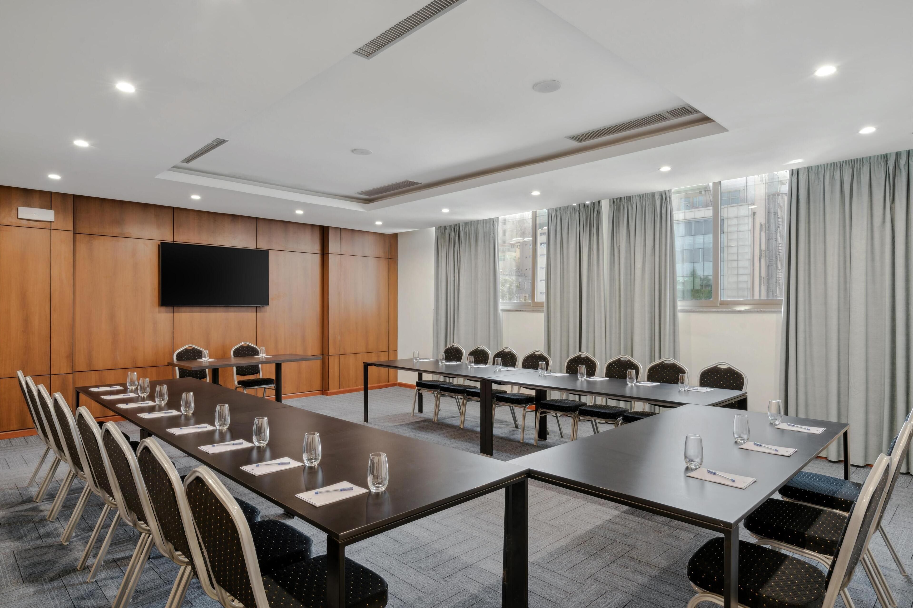 A conference room with a long table, chairs, a flat-screen TV, and large windows with curtains.