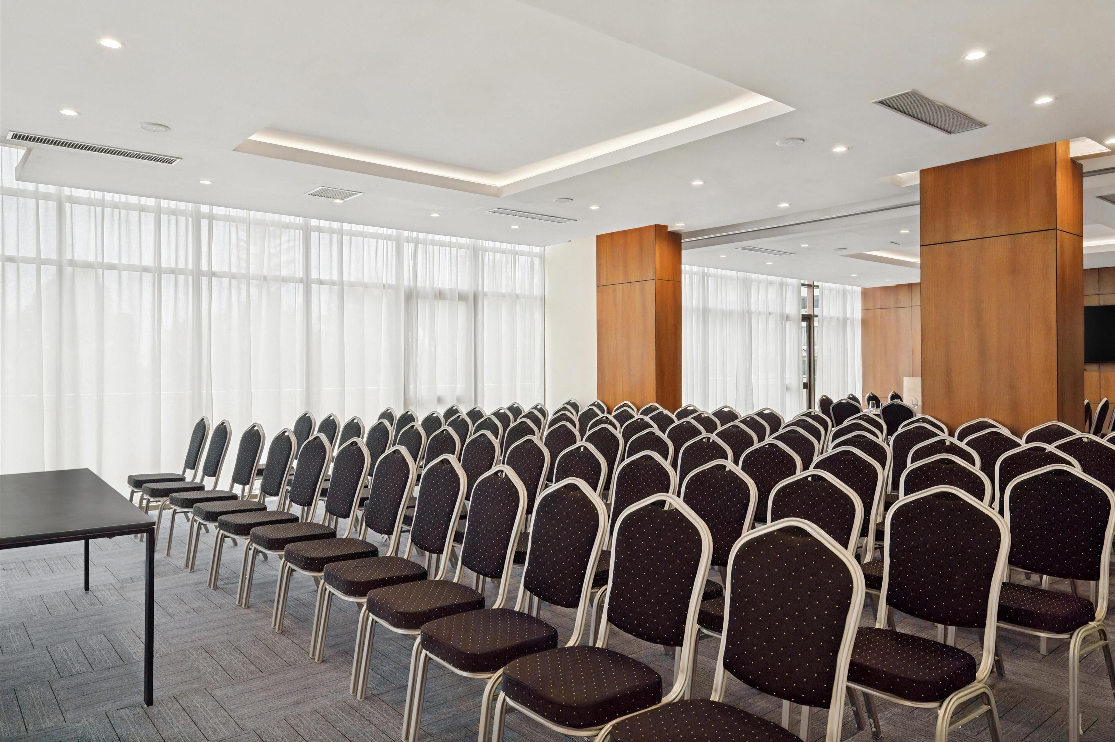 A conference room with rows of chairs, a long table, and large windows with sheer curtains.