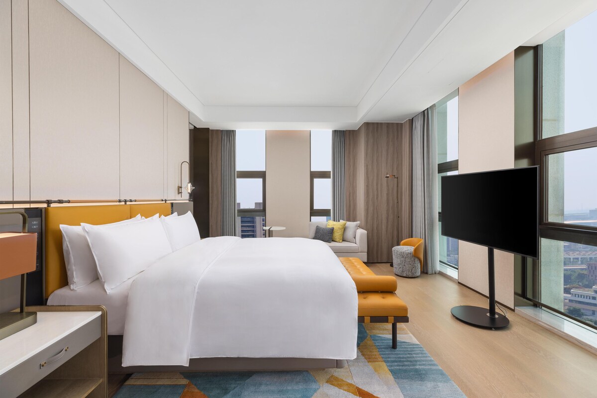 A hotel room with a bed, a TV, a sofa, a small table, and a large window with a city view.