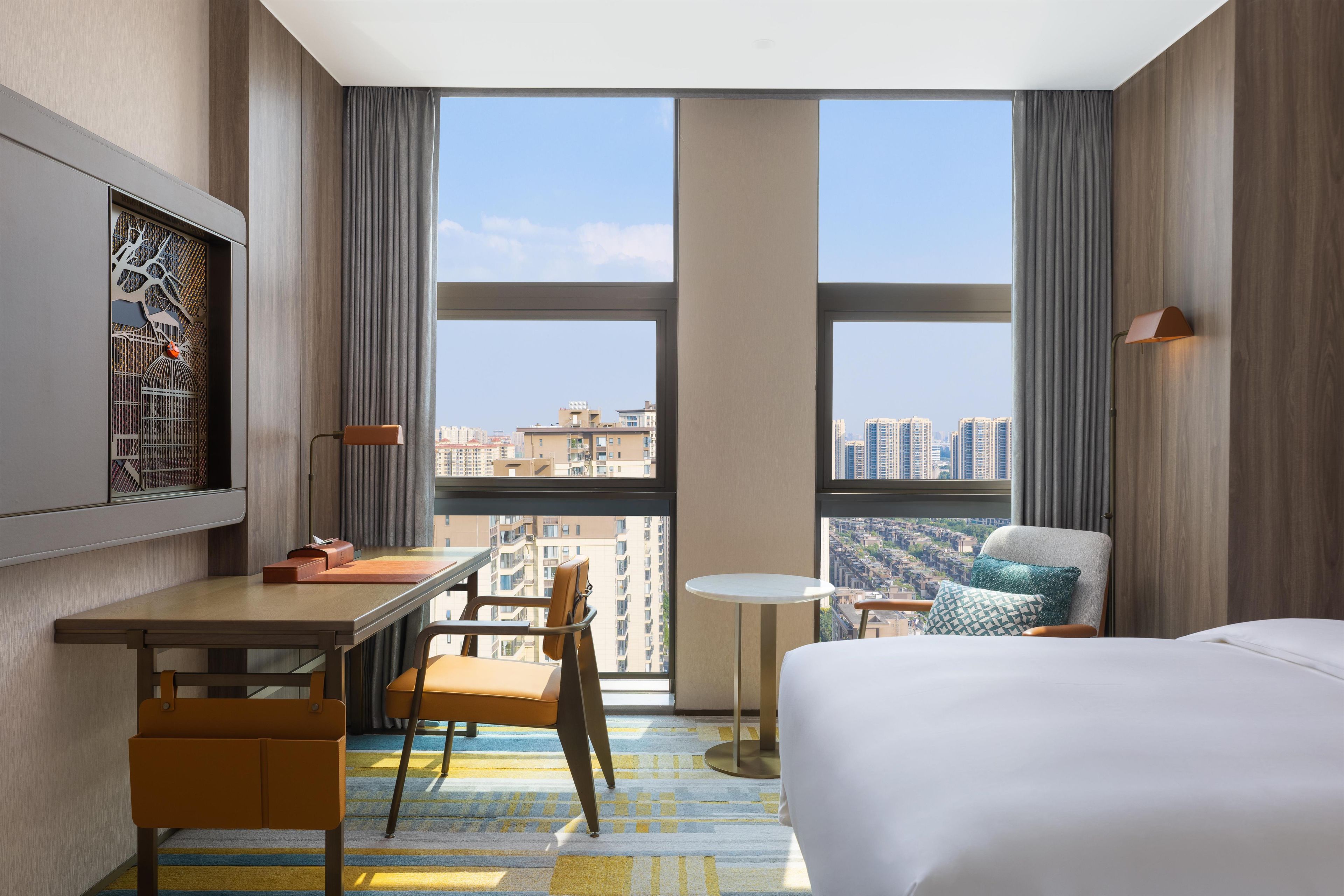 A hotel room with a bed, a desk, a chair, a small table, and a view of the cityscape through large windows.