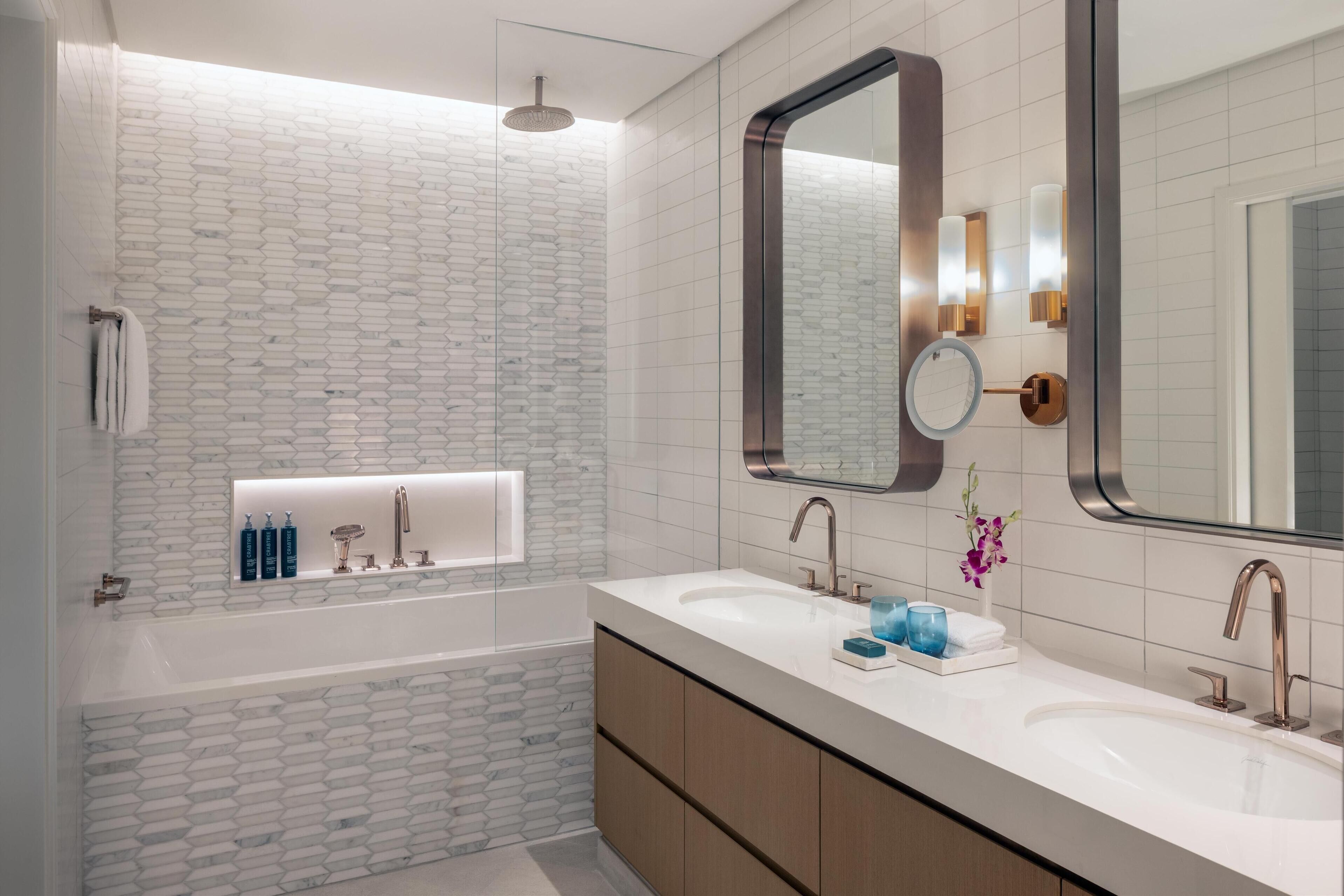 Une salle de bain moderne avec un mur aux motifs blancs et gris, un grand miroir, une baignoire et un meuble-lavabo doté d’un évier et de deux robinets.
