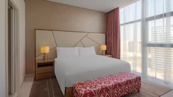 Habitación de hotel con una cama grande, dos mesitas de noche con lámparas, un banco estampado y vistas a edificios a través de cortinas traslúcidas.