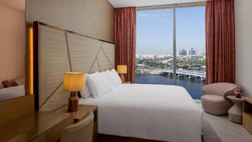 A hotel room with a large bed, a desk, a chair, and a view of a cityscape through a large window.