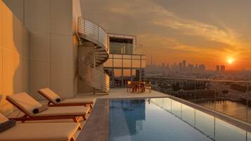 A rooftop infinity pool with lounge chairs, a spiral staircase, and a city skyline view at sunset.