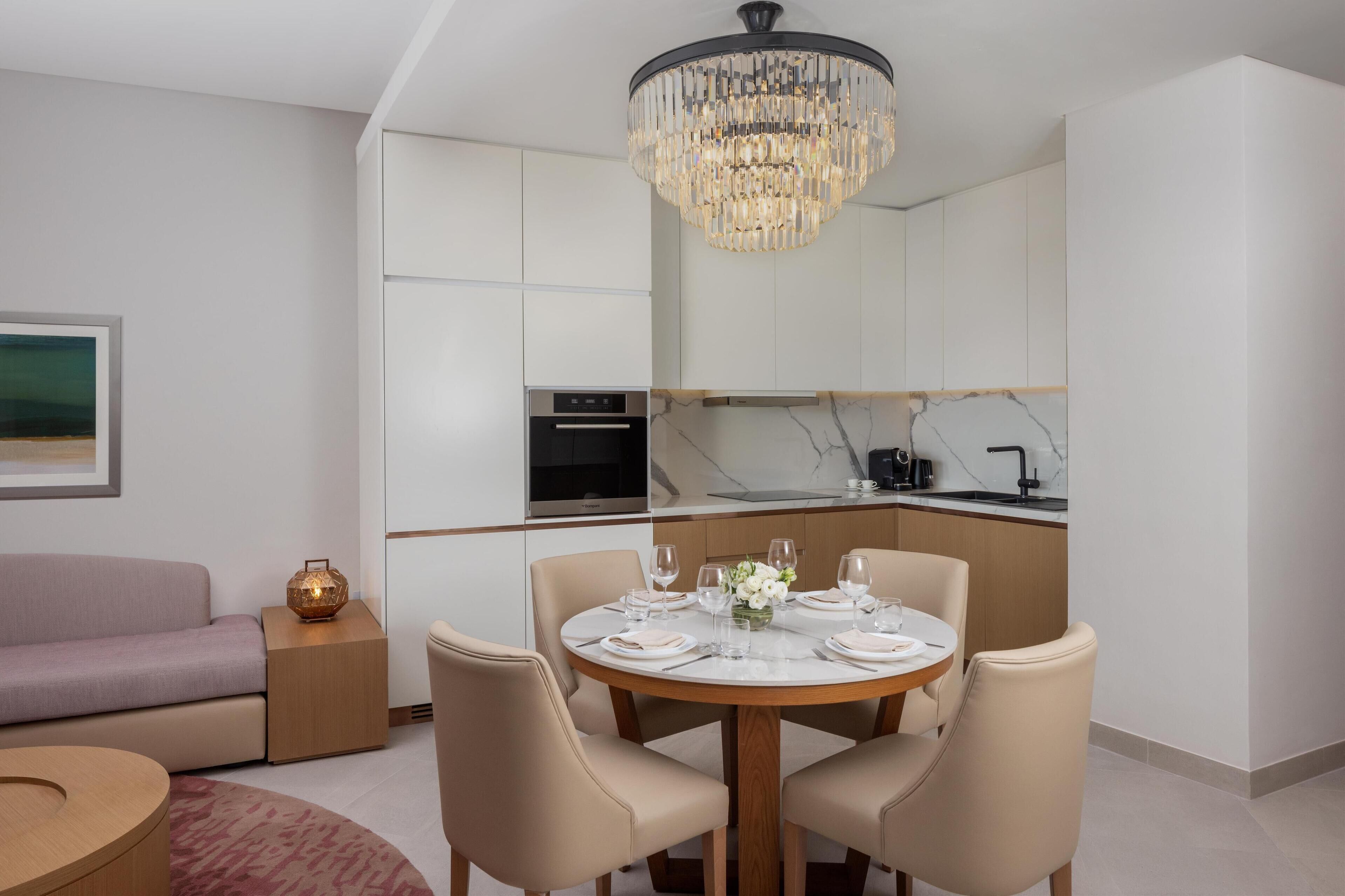 Un espace de salle à manger moderne avec une table ronde dressée pour quatre personnes, un lustre et une cuisine en arrière-plan.
