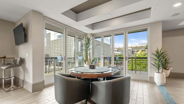 Un salon moderne avec une table ronde, deux chaises, un téléviseur fixé au mur et de grandes portes-fenêtres coulissantes donnant sur un balcon aménagé avec du mobilier d’extérieur.