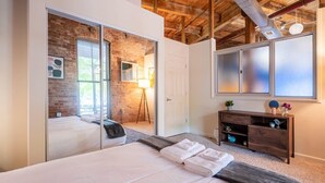 A bedroom with a large bed, a wooden dresser, a mirror, and a brick wall. - 100 Year-Old Historic Brick 2BR Loft -Large Patio1 (Durham)